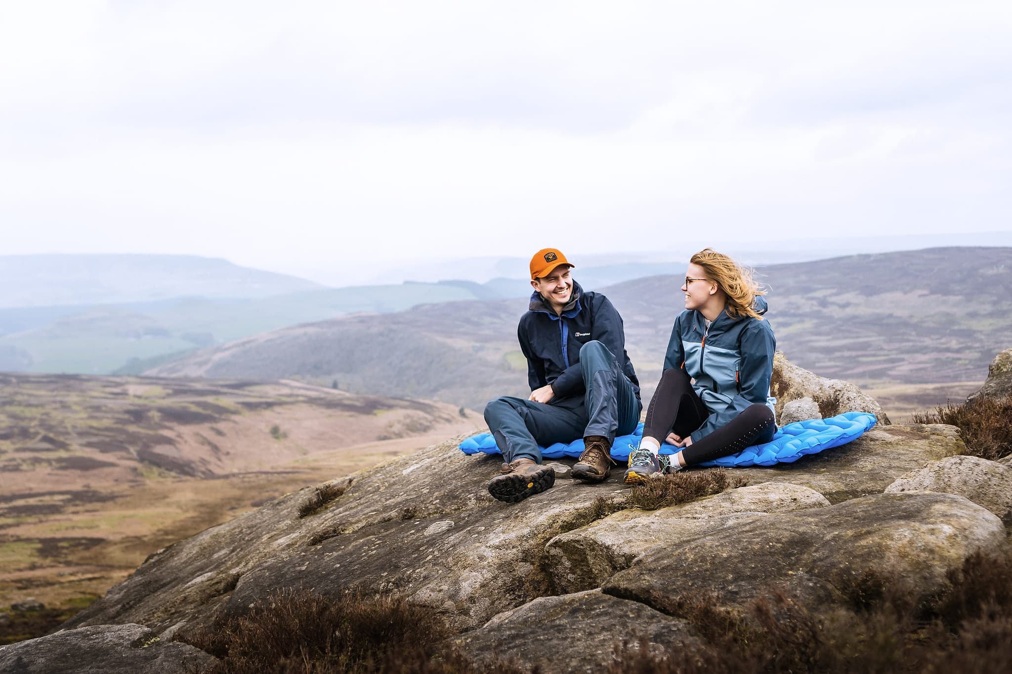 Two wild campers sat on a camping mat on a gritstone edge above a misty UK valley